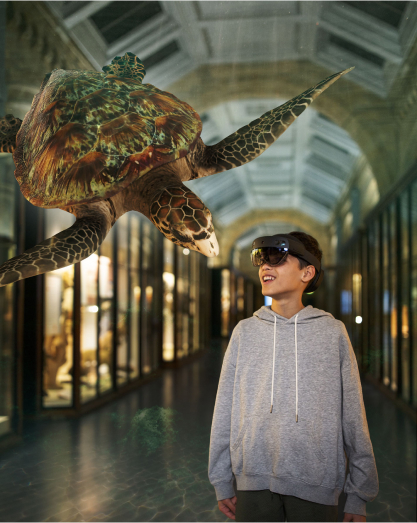 A young boy wearing a gray hoodie and VR headset looks at a virtual sea turtle in a museum hallway.