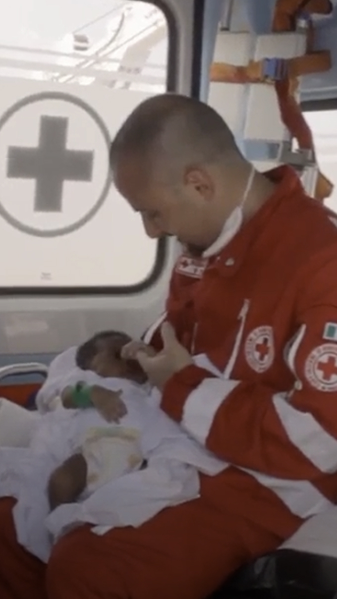 A paramedic in a red uniform feeds an infant inside an ambulance with a Red Cross symbol visible on the window.